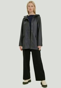 Casaco de chuva preto impermeável com capuz, botões de pressão, bolsos laterais e um corte descontraído, usado sobre calças largas pretas e sapatos bege.