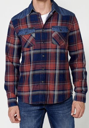 Homme portant une chemise à carreaux rouge et bleu marine à manches longues avec des poches poitrine boutonnées et un jean en denim bleu.
