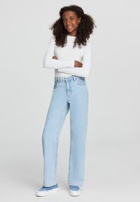 Niebieskie spodnie z wysokim stanem, proste, w jasnym odcieniu, połączone z białą bluzką z długim rękawem i niebieskimi trampkami. Gładka tekstura, minimalne detale.