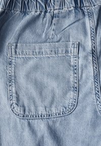 Hellblauer Denim-Stoff mit einem gerafften Bund, der eine quadratische aufgesetzte Tasche aufweist. Die Nähte sind entlang der Kanten sichtbar. Glatte Textur.
