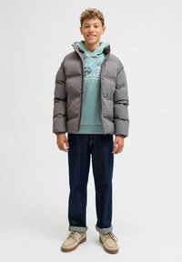 Chaqueta de plumas gris con capucha, llevada sobre un sudadera verde claro y jeans azules. Tela texturizada con costuras visibles y cierre de cremallera.