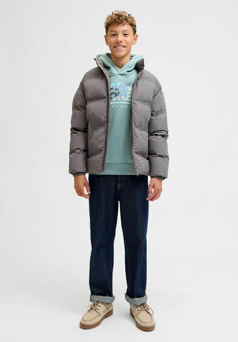 Chaqueta de plumas gris con capucha, llevada sobre un sudadera verde claro y jeans azules. Tela texturizada con costuras visibles y cierre de cremallera.