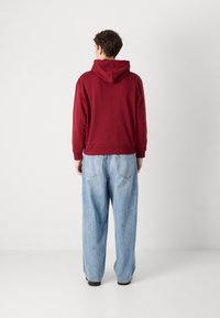 Felpa con cappuccio bordeaux dal fit rilassato, con polsini e orlo a coste, abbinata a jeans a gamba larga di un blu chiaro. Il tessuto sembra morbido.