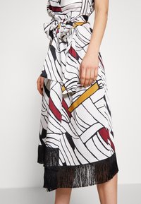 Jupe blanche avec des lignes noires audacieuses et des motifs géométriques rouges et jaunes. Comprend un ourlet noir à franges et un détail de taille nouée.