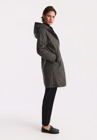 Pikowana zielona parka z kapturem, z bocznymi kieszeniami i gładką teksturą. Nosi pod spód czarne spodnie i czarne buty wsuwane.