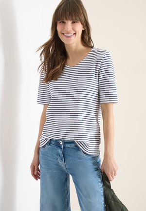 Jeune femme souriante, portant un t-shirt à manches courtes rayé blanc et bleu marine, un jean bleu, tenant un sac à main matelassé vert foncé.