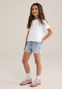 Weißes Baumwoll-T-Shirt mit einem kleinen Sonnenmotiv, kombiniert mit hellblauen Jeansshorts mit fransigen Säumen. Rosa und graue Sneakers mit weißen Schnürsenkeln.
