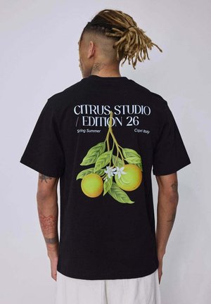 Homme aux dreadlocks vu de dos portant un t-shirt noir avec un motif de fruits agrumes et feuilles et le texte « Citrus Studio Edition 26 Spring Summer Capri Italy. »