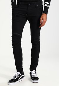 Svarta slim-fit jeans med dragkedjedetaljer vid knäna, tillverkade av stretchigt material. Bärs tillsammans med svarta högstövlade sneakers med vita sulor och snören.