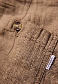 Brunt herrbet fabric med en synlig knapp och en vit etikett märkt "Lindbergh." Nära bild av textur och sömnadsdetaljer.