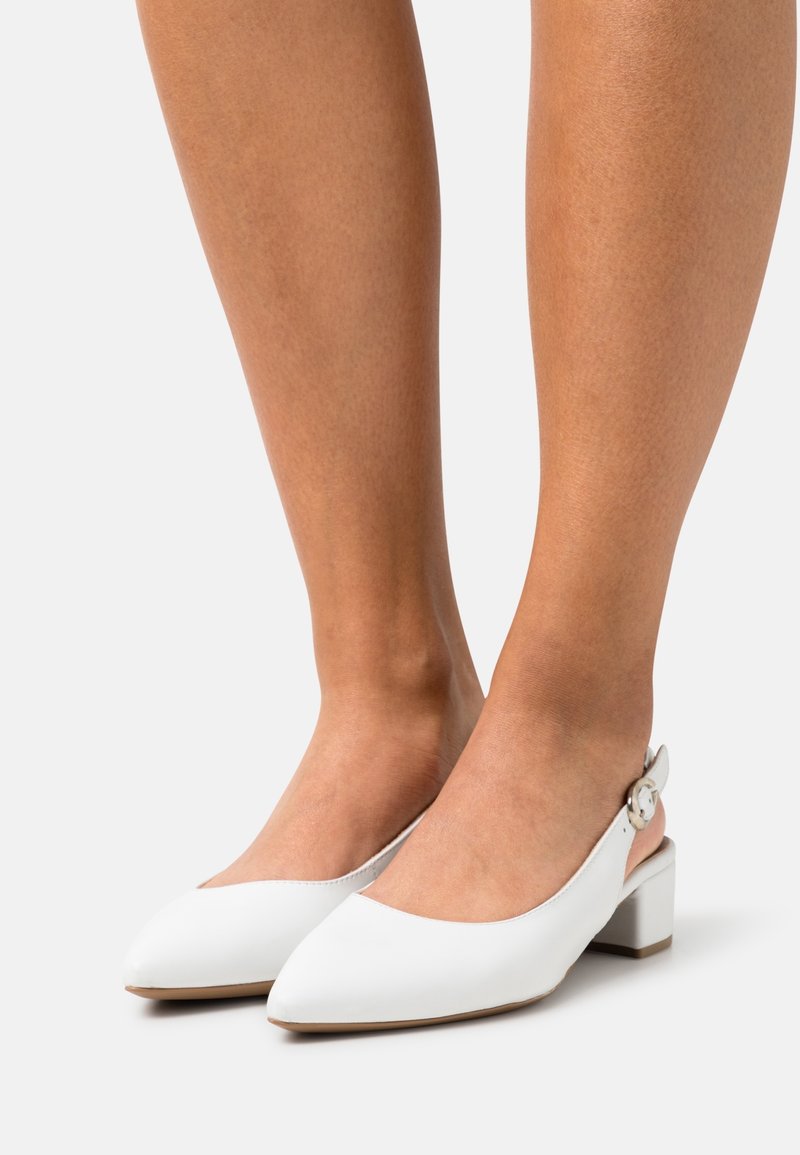 Chaussures slingback en cuir blanc avec bouts pointus et petits talons blocs portées pieds nus sur un fond clair.