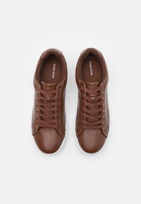 Baskets en cuir marron avec bout arrondi, semelles blanches et lacets marron. L'intérieur présente un doublage texturé marron avec le nom de la marque.