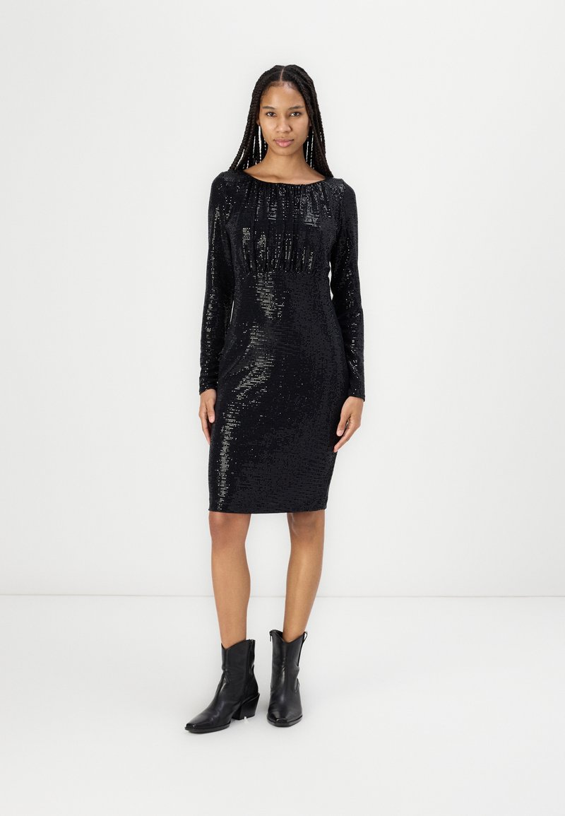 Femme portant une robe noire à manches longues, en sequins, de longueur genou avec des bottines noires, se tenant contre un fond clair.