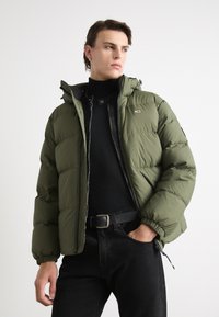 Puffige olivgrüne Jacke mit Kapuze, Reißverschluss und elastischen Bündchen, getragen über einem schwarzen Rollkragenpullover und schwarzen Jeans.