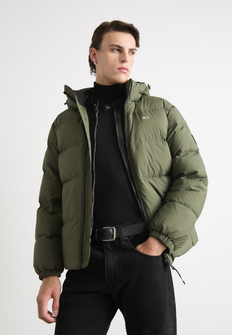 Puffige olivgrüne Jacke mit Kapuze, Reißverschluss und elastischen Bündchen, getragen über einem schwarzen Rollkragenpullover und schwarzen Jeans.