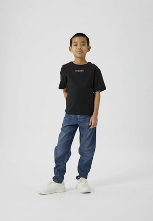 Svart bomullstrøye med "Studio edition" trykket i hvitt, kombinert med medium blå jeans og hvite sneakers, satt mot en enkel bakgrunn.