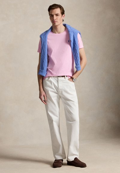 Ung mand iført en pink T-shirt, hvide bukser, brune loafers og en blå sweater, der er draperet over skuldrene, mod en neutral baggrund.