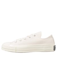 Sneakers in tela a bassa altezza di color beige chiaro con suola in gomma bianca, sei occhielli in metallo e una patch con logo vicino al tallone.