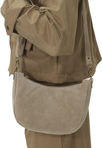 Beige Wildleder-Crossbody-Tasche mit geschwungener Form, Reißverschluss und verstellbarem Tragegurt, getragen über einem braunen Outfit.