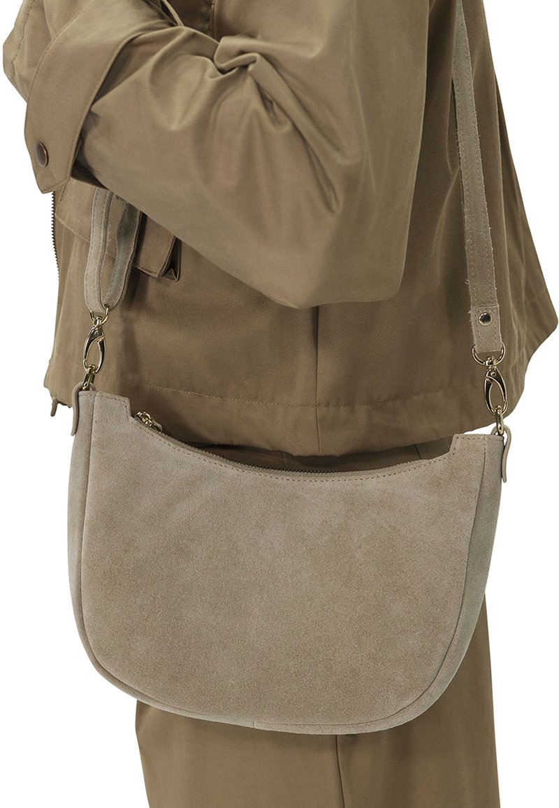 Beige Wildleder-Crossbody-Tasche mit geschwungener Form, Reißverschluss und verstellbarem Tragegurt, getragen über einem braunen Outfit.