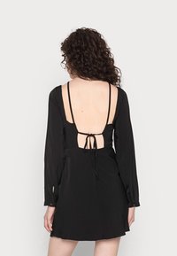 Vestido preto com mangas longas, costas abertas e detalhe de laço ajustável. Tecido suave com um corte relaxado e ligeira abertura na bainha.