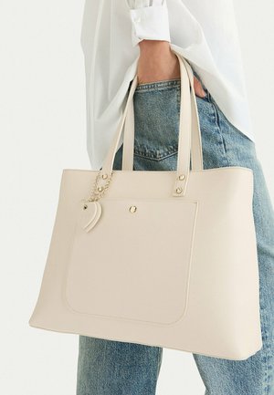 Persona con jeans azules y camisa blanca sostiene un bolso tote grande beige con bolsillo frontal y un colgante en forma de corazón.