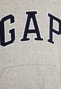 Felpa grigia con logo audace "GAP" in blu navy. Tessuto morbido con una finitura testurizzata. Include una tasca a marsupio frontale.