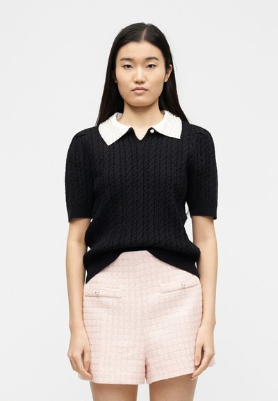 Jeune femme portant un pull à manches courtes noir texturé avec un col blanc orné et un short taille haute rose clair avec des poches devant.