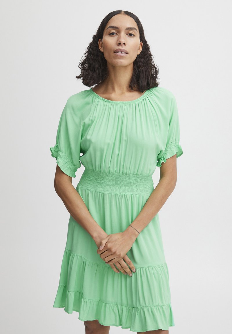 Grünes, knielanges Kleid mit gerafftem Bund, kurzen Puffärmeln und Rüschensaum. Aus einem fließenden, leichten Stoff gefertigt.