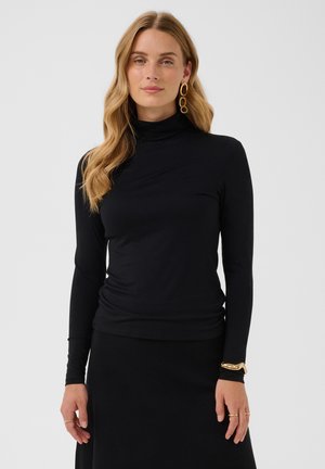 Femme aux longs cheveux blonds portant un col roulé noir et une jupe, des boucles d'oreilles créoles dorées, un bracelet en or et des bagues sur un fond gris uni.