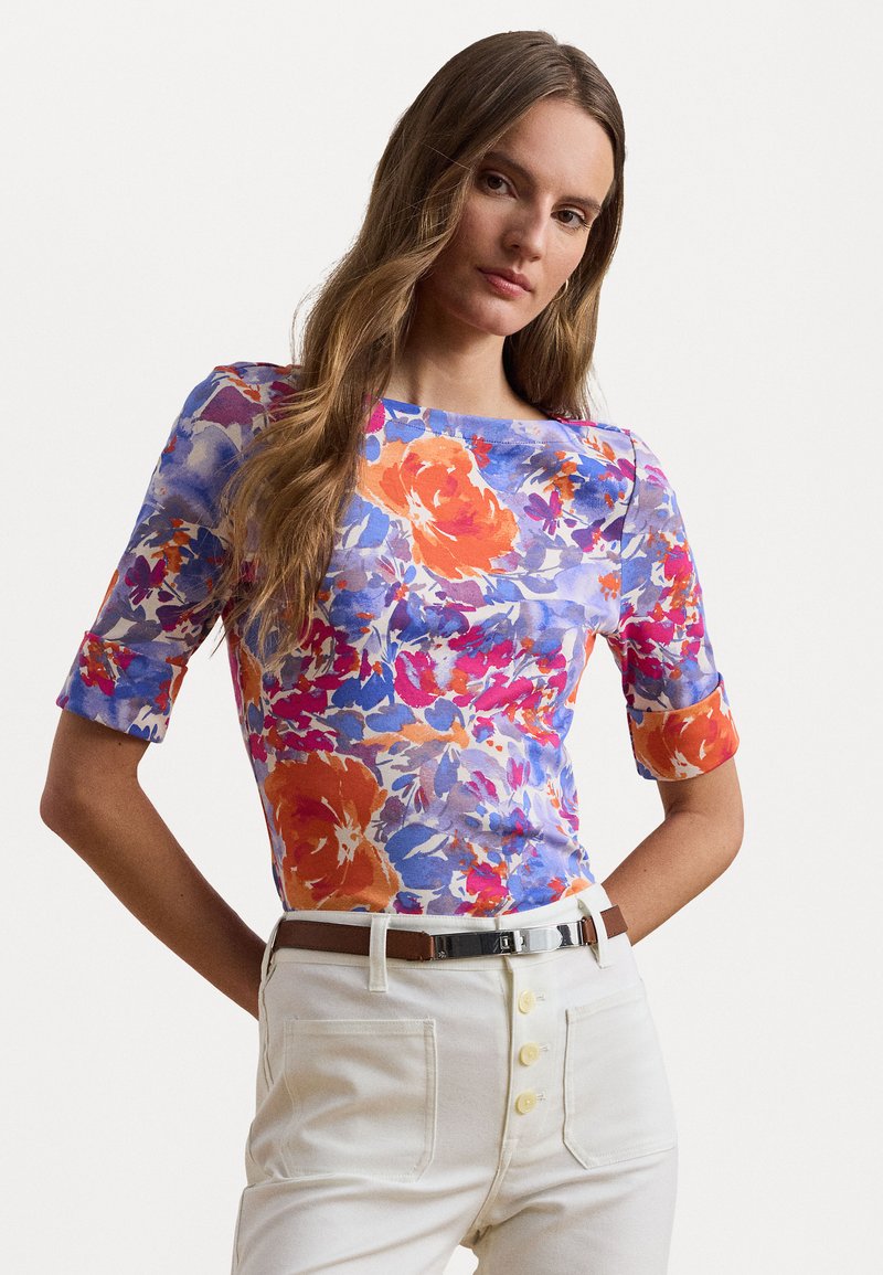 Top floral dans des tons de bleu, orange et rose ; manches courtes avec poignets roulés ; design ajusté ; porté avec un pantalon crème et une ceinture marron.