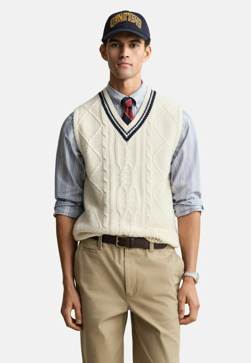 Nuori mies, jolla on tummansininen lakki, vaaleanbeige villapaita, jossa on joustokkaita, raidallinen paita, jonka hihat on käärityt, raidallinen solmio, vaaleat housut ja rannekello.