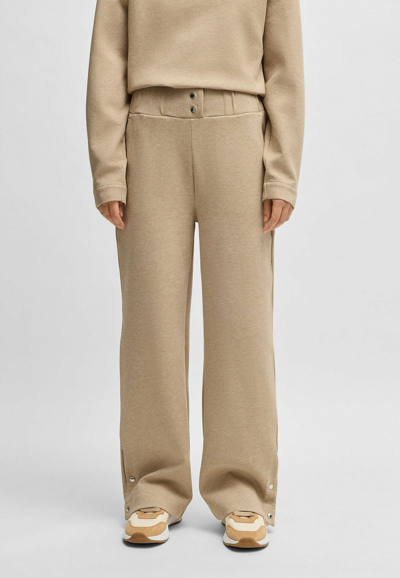 Beige, leveled sweatpants dengan pinggang rib yang lebar, dua kancing depan, dan detail snap di pergelangan kaki. Tekstur kain yang halus.