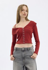 Red ribbed crop top with long sleeves, off-the-shoulder design, large collar, and metallic button accents down the front; paired with faded gray jeans.