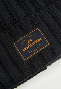 Czarna dzianinowa czapka beanie o prążkowanej teksturze. Posiada prostokątną metkę z pomarańczowym napisem "AUTHENTIC COALITION WORKWEAR" na granatowym tle.