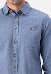 Camisa de franela azul con un bolsillo en el pecho, cierre frontal de botones y botones de madera. La tela tiene una textura suave y un diseño casual.