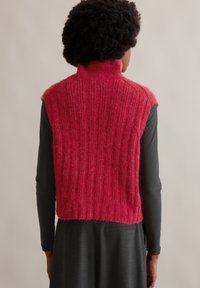 Roter ärmelloser Pullover mit hohem Kragen, der eine vertikale Rippstruktur und weiches, flauschiges Material aufweist. Wird über einem schwarzen Langarmshirt getragen.