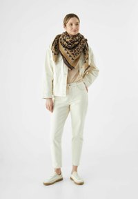 Beige denimjak over een beige top, gecombineerd met witte broek en witte sneakers; een grote bruin-zwarte luipaardprint sjaal maakt de look compleet.