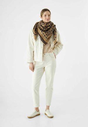 Veste en denim beige sur un top beige, associée à un pantalon blanc et des baskets blanches ; une grande écharpe à imprimé léopard brun et noir vient compléter le look.