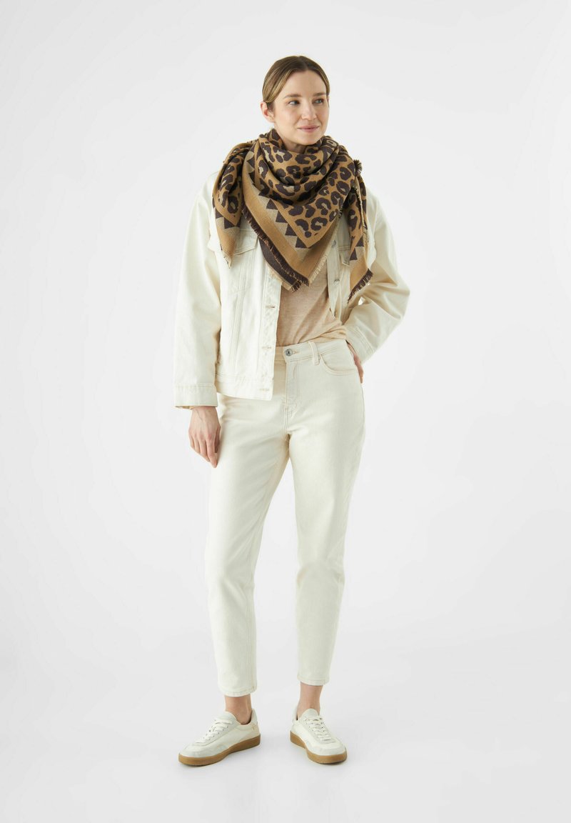 Beige denimjak over een beige top, gecombineerd met witte broek en witte sneakers; een grote bruin-zwarte luipaardprint sjaal maakt de look compleet.
