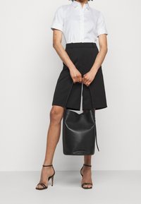 Chemise blanche à manches courtes avec boutons, short noir ajusté, talons noirs à brides et grand sac cabas en cuir noir.