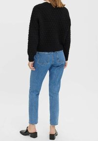 Schwarzer Strukturpullover mit rundem Ausschnitt, kombiniert mit hellblauen hochgeschnittenen Jeans. Die Schuhe sind schwarz mit einem Blockabsatz.