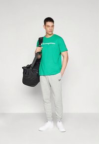 Zelené bavlněné tričko s logem "Champion", šedé tepláky a bílé tenisky. Model drží černou tašku přes rameno s popruhem.