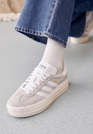 Stopa w białych butach sportowych Adidas Gazelle, białych skarpetkach i niebieskich dżinsach na jasno szarej, nakrapianej podłodze.