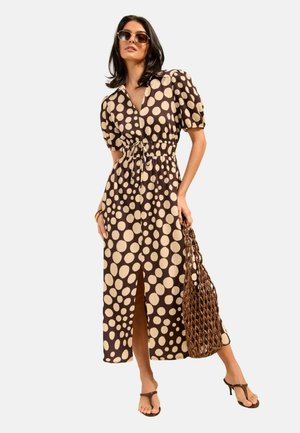 Kobieta w brązowej i beżowej midi sukience w groszki z bufkami na rękawach, trzyma plecioną torbę, nosi okulary przeciwsłoneczne i brązowe sandały na obcasie.