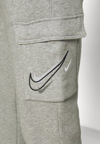 Šedé tepláky s kapsou, na které jsou umístěny čtyři překrývající se loga Nike swoosh v černé, bílé, šedé a tmavě šedé barvě po straně.