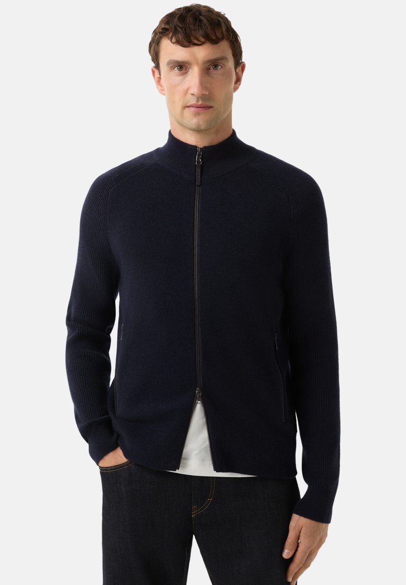 Námořnický svetr na zip s žebrovanou texturou, stojáčkovým límcem a bočními kapsami, vrstvený přes bílou košili, v kombinaci s tmavými džínami.