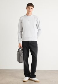 Felpa grigia chiara con scollo rotondo e logo in rilievo, abbinata a jeans neri. Il modello tiene in mano una borsa grigia e indossa sneakers bianche.