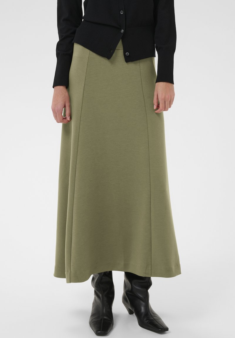 Falda larga color verde oliva en tejido de punto suave, con una silueta fluida y detalles de costuras verticales, combinada con botas negras de tacón.