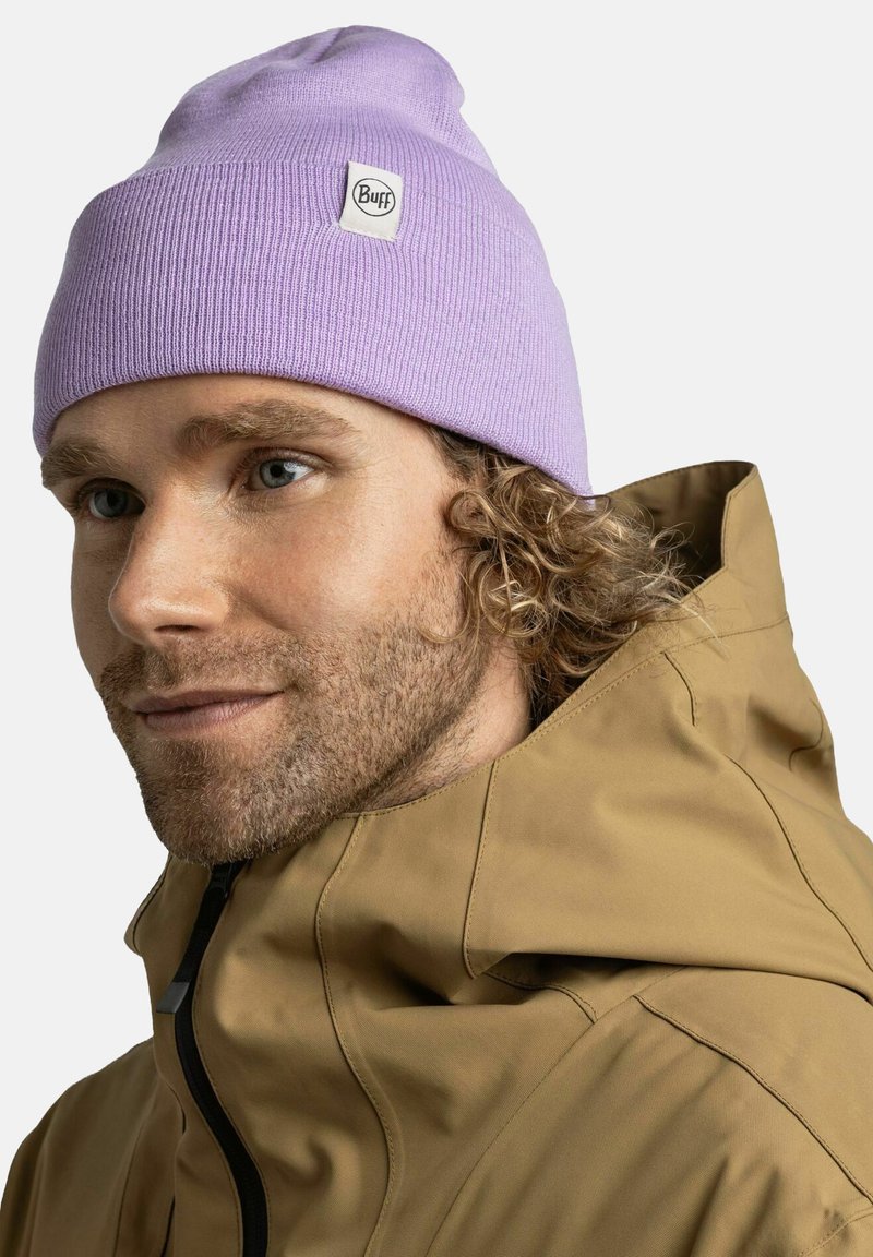 Lavender knit beanie with a logo patch, worn by a person with curly hair, paired with a beige jacket featuring a high collar and zipper.